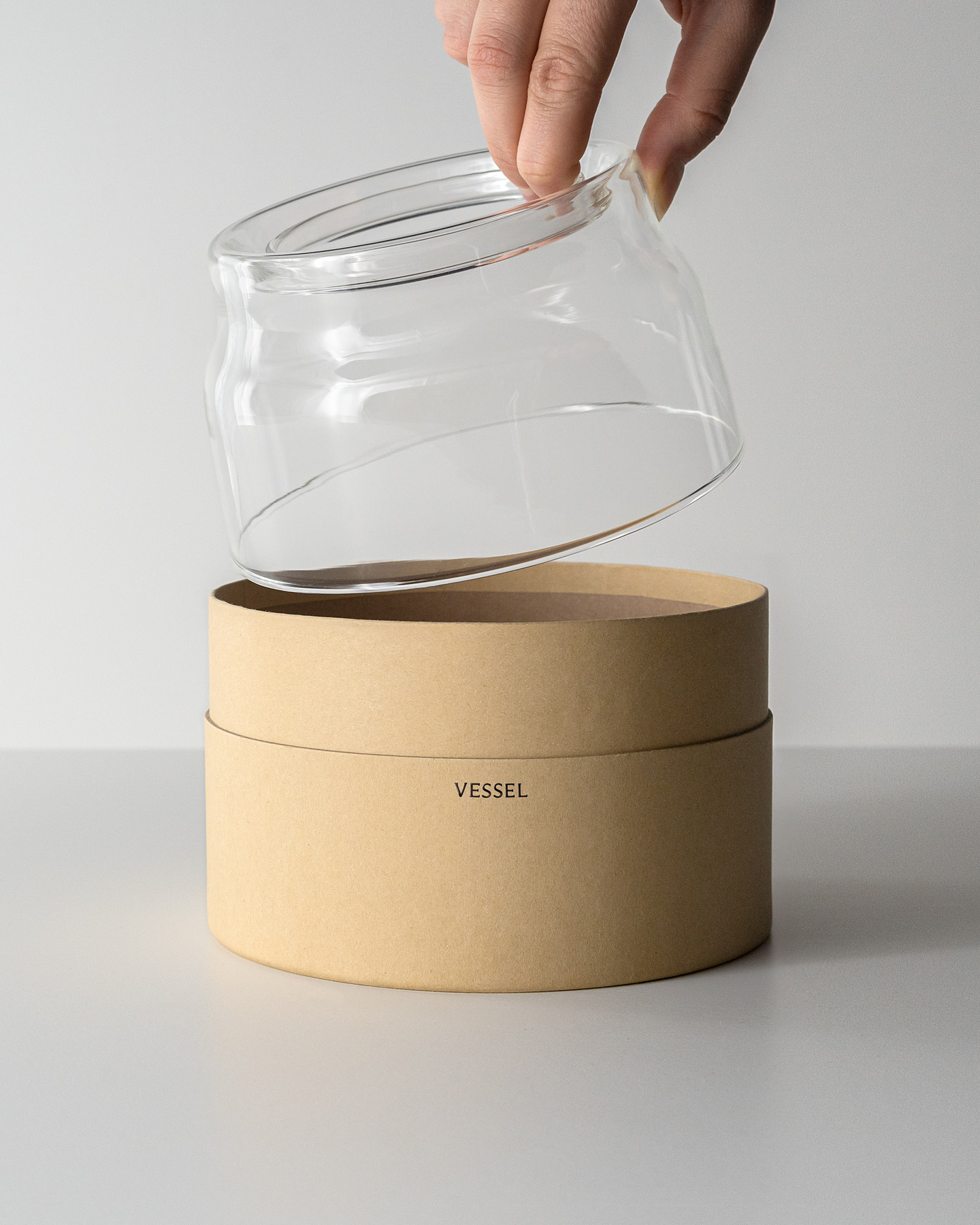 Hand lifting a clear glass vessel over a round cardboard packaging box