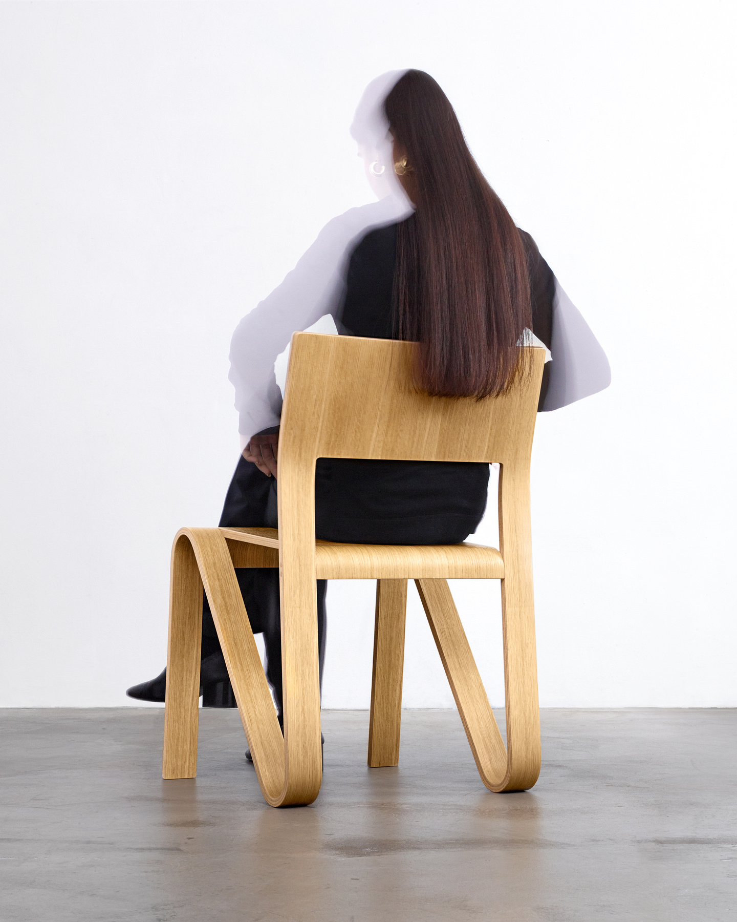 Side perspective of a person seated on a light wooden chair against a plain wall.