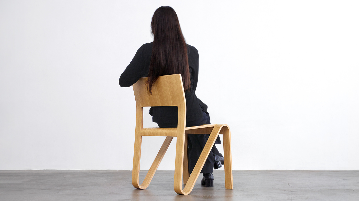 Back view of a person seated on a light wooden chair in a studio setting.