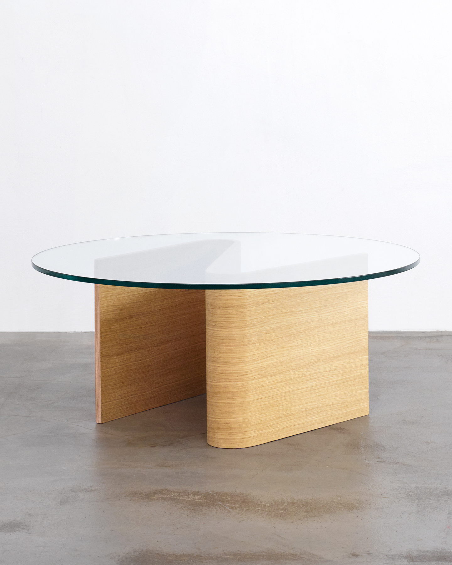 Circular glass coffee table resting on a curved wooden base on a concrete floor.