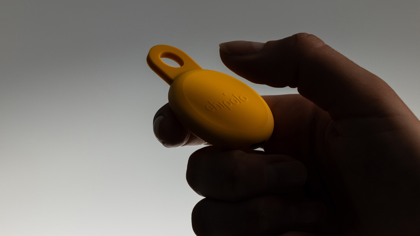 Hand holding a small yellow Chipolo Loop tracker against a grey background.