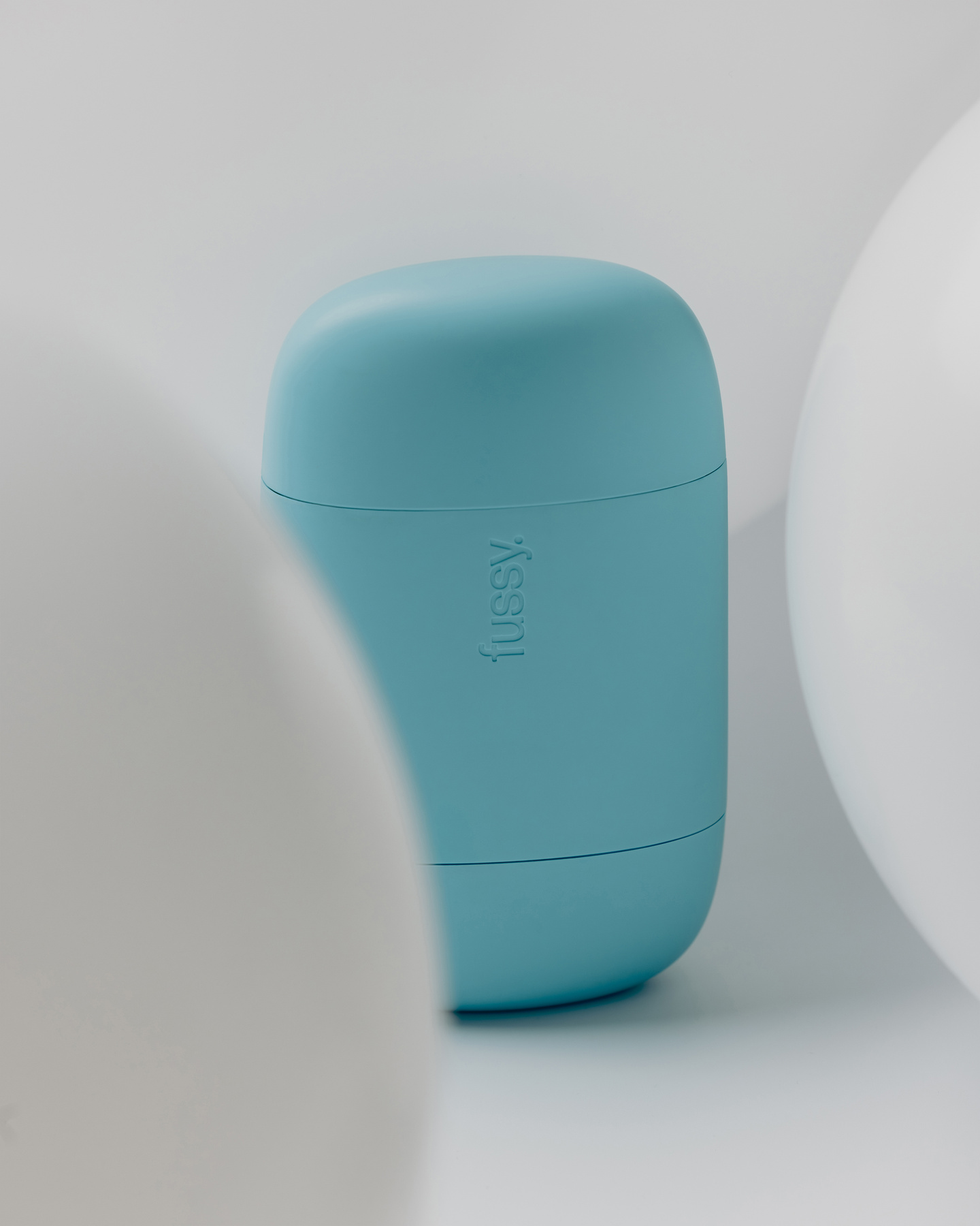 Small turquoise Fussy deodorant case set between soft white spherical shapes.