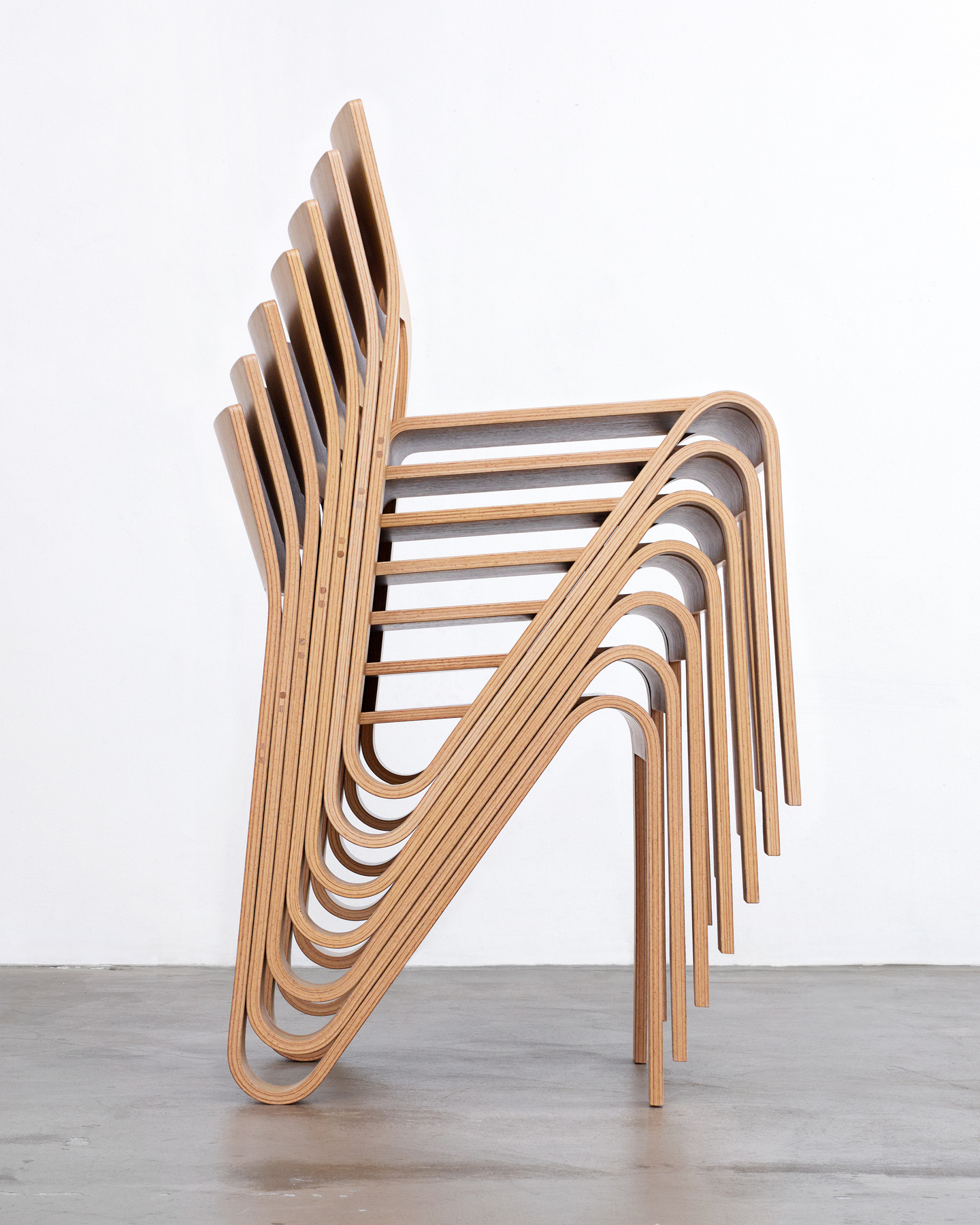 Side studio view of a pile of light wooden chair on a concrete floor.