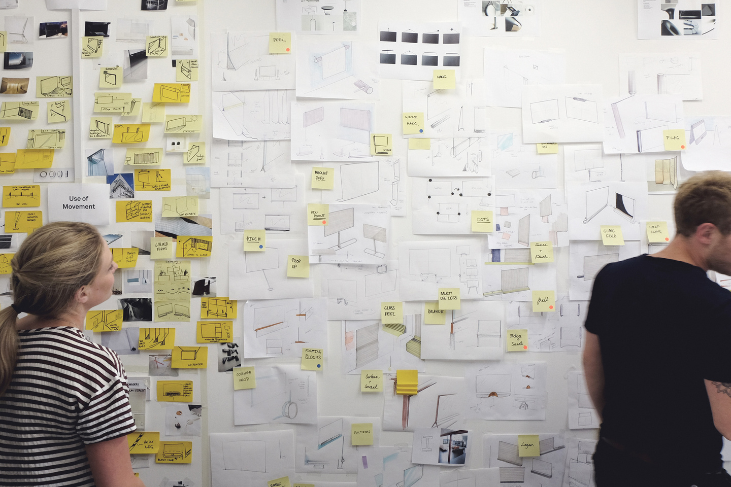 Designers reviewing sticky notes and concept sketches on a whiteboard wall.