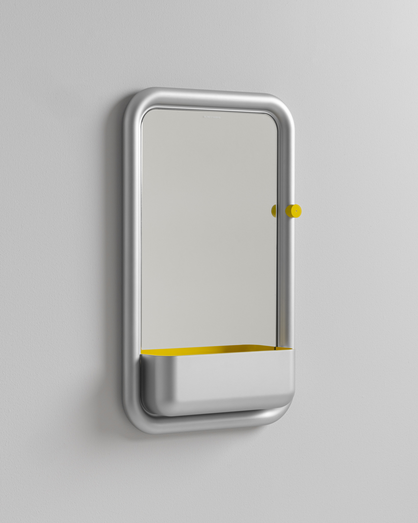 Wall-mounted rectangular mirror unit with a yellow dial dial accent and lower tray on a light wall