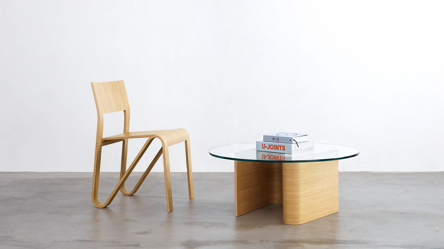 Light wooden chair standing next to a circular glass-topped coffee table in a studio space.
