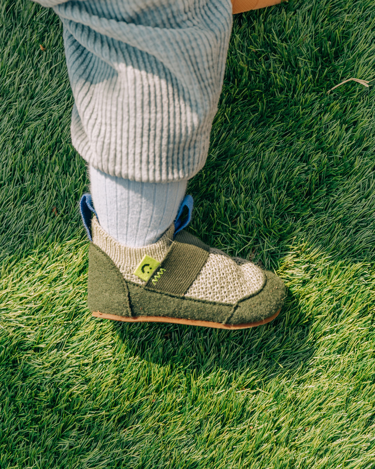 Child wearing a green fabric shoe standing on green lawn grass outdoors.