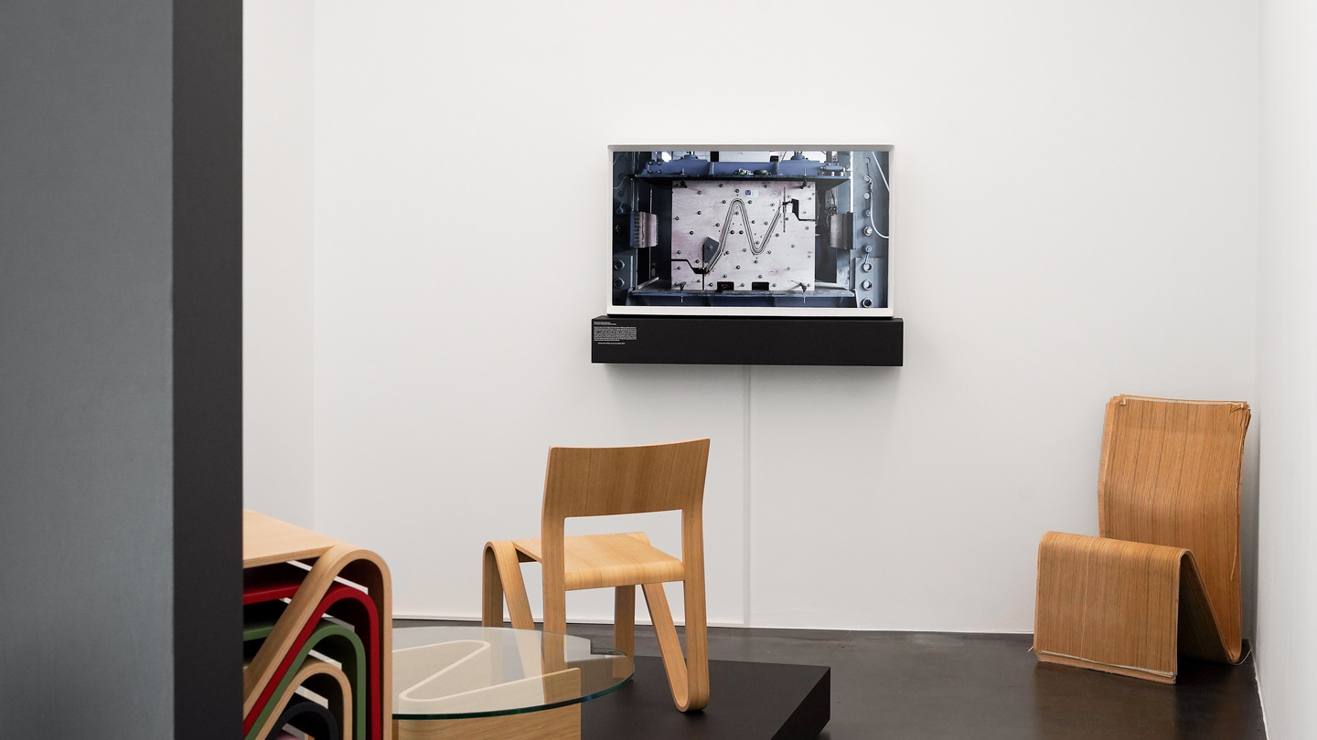 Interior of an exhibition space with a wall-mounted screen, wooden seating and artwork.